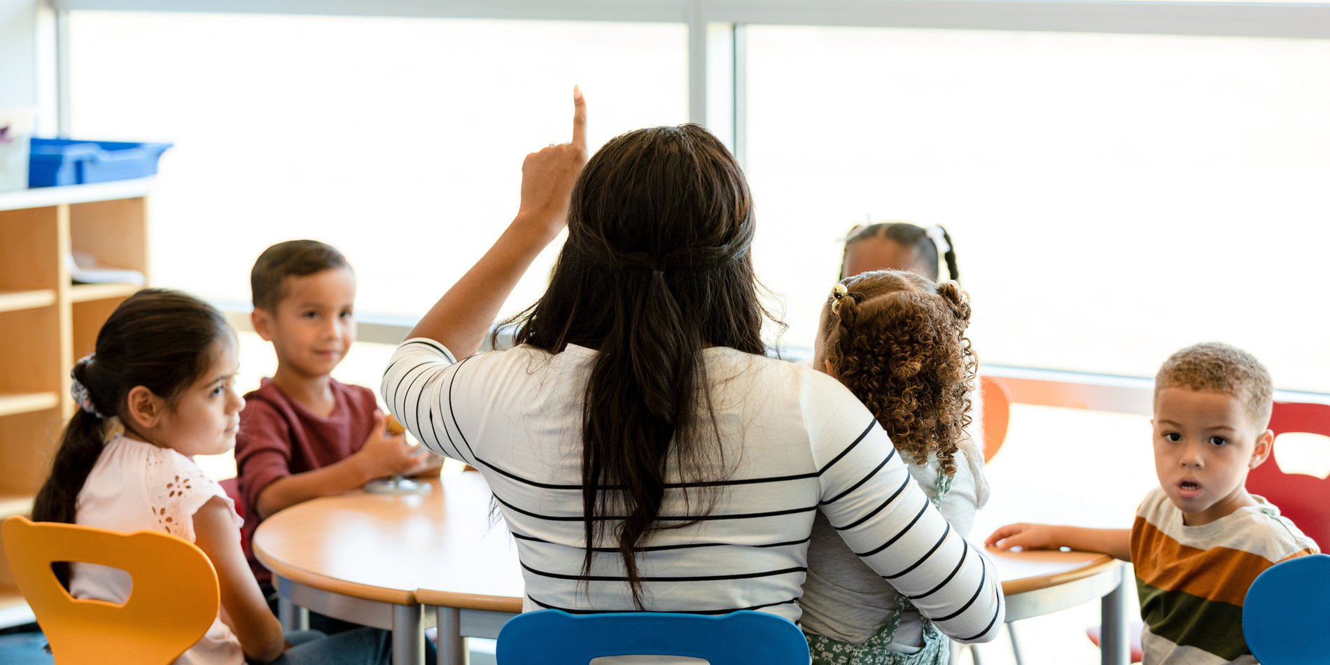 Young students sit at a table with a teacher in a classroom near sunlit windows