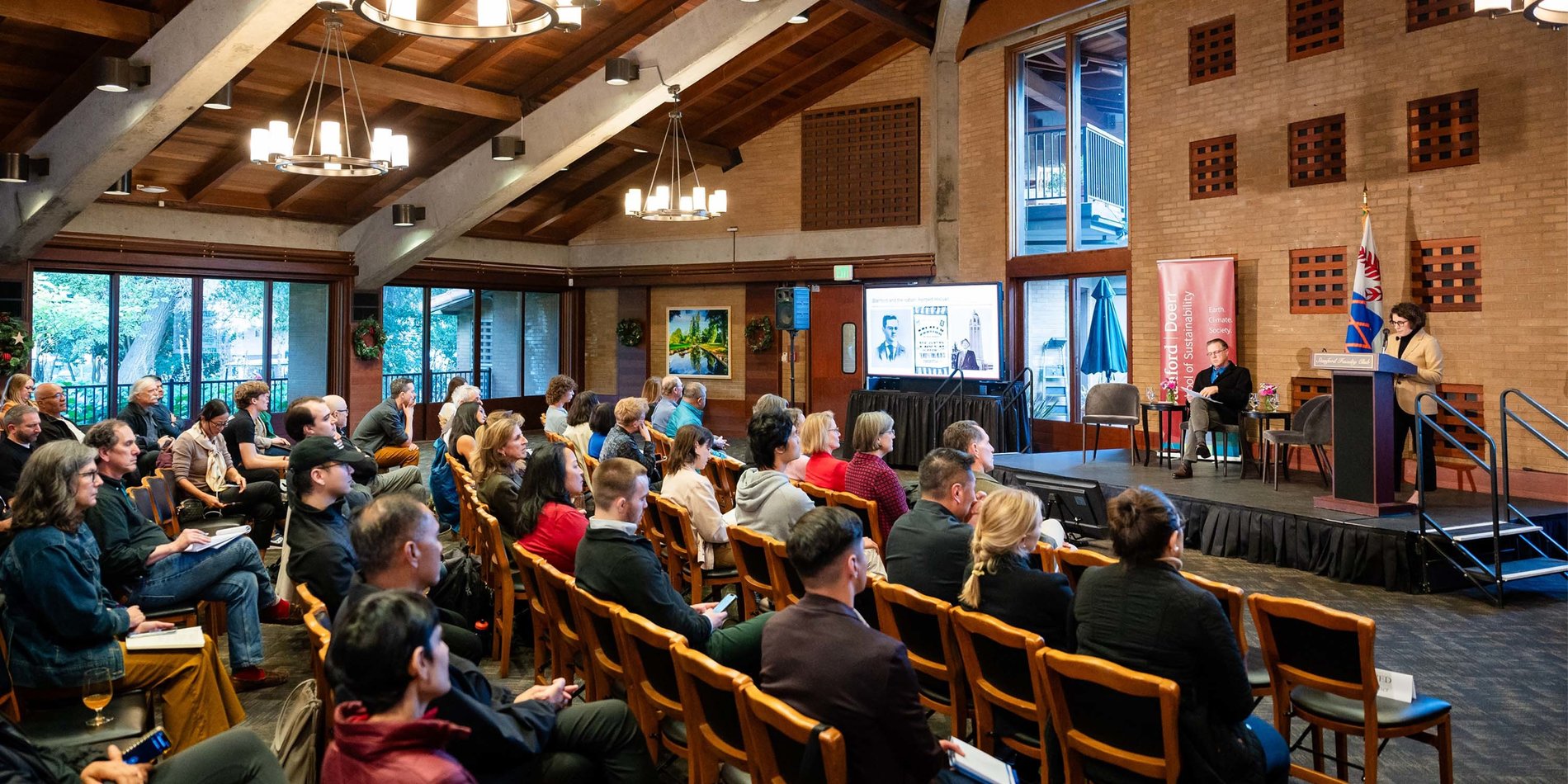 Wide view of event room with Emily Levine speaking at the podium and Mitchell Stevens sitting on stage