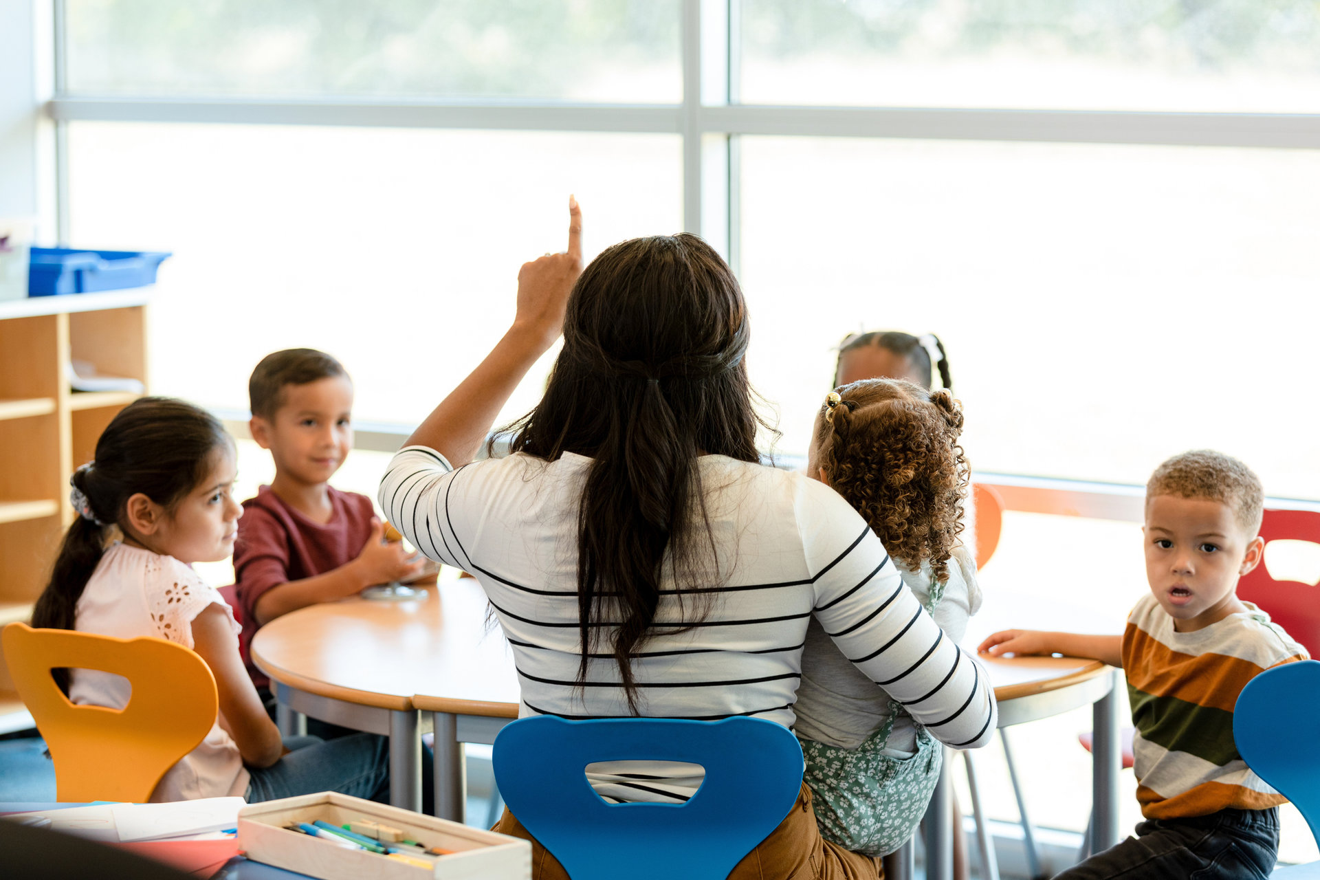 Young students sit at a table with a teacher in a classroom near sunlit windows