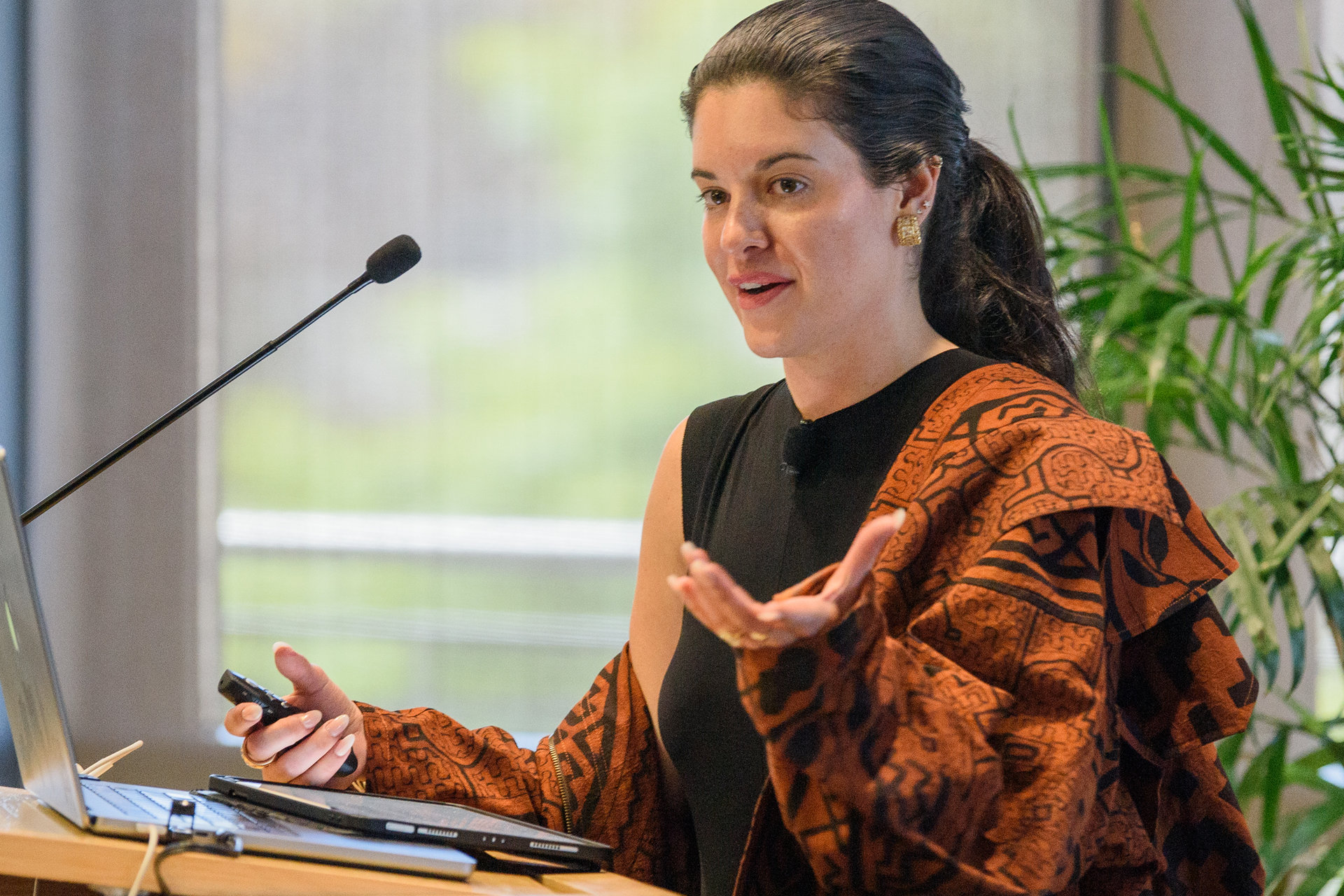 A woman wearing a rust-colored jacket with a geometric pattern gestures as she speaks into a microphone at a podium