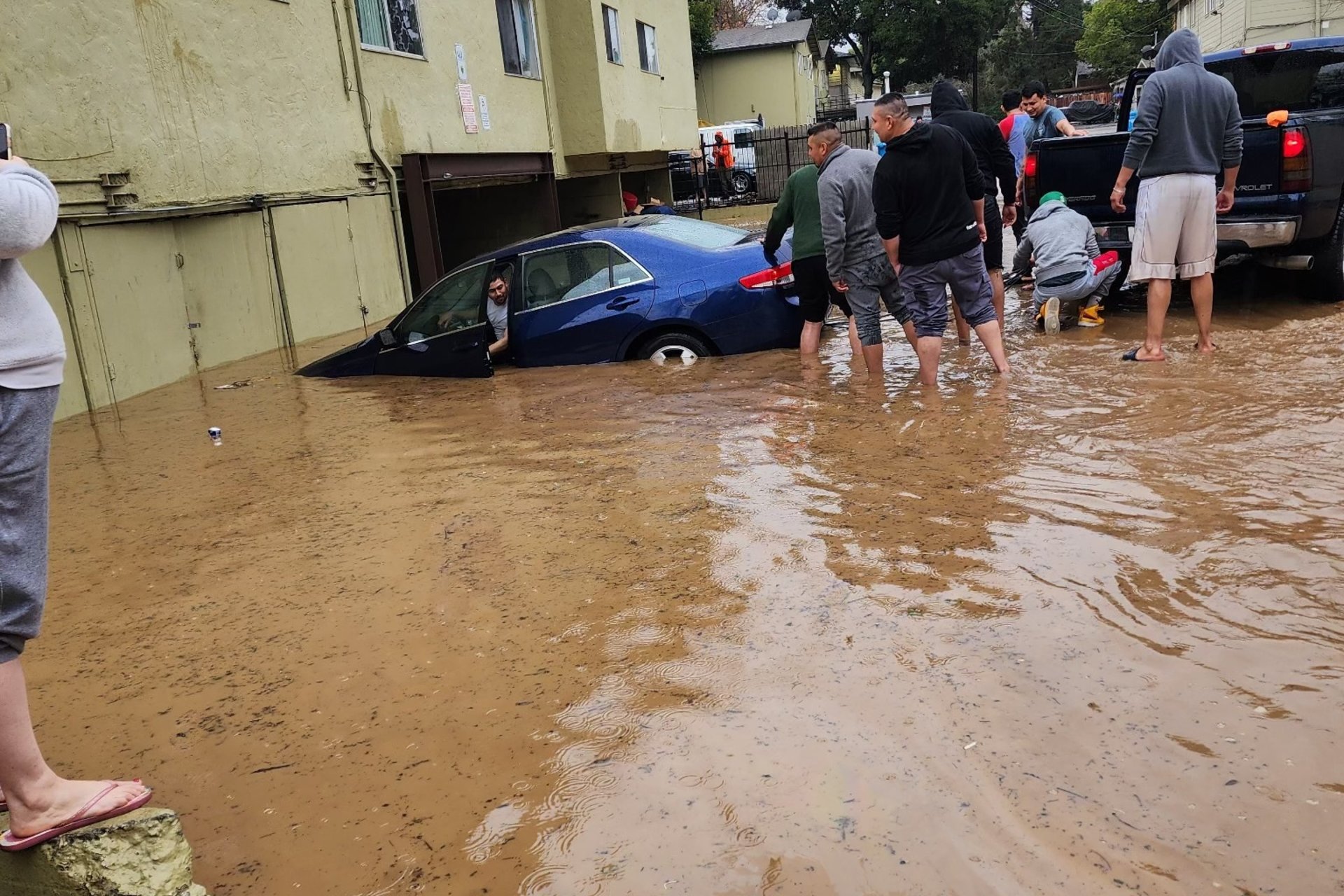 People wade in brown floodwater to help a neighbor near a submerged car