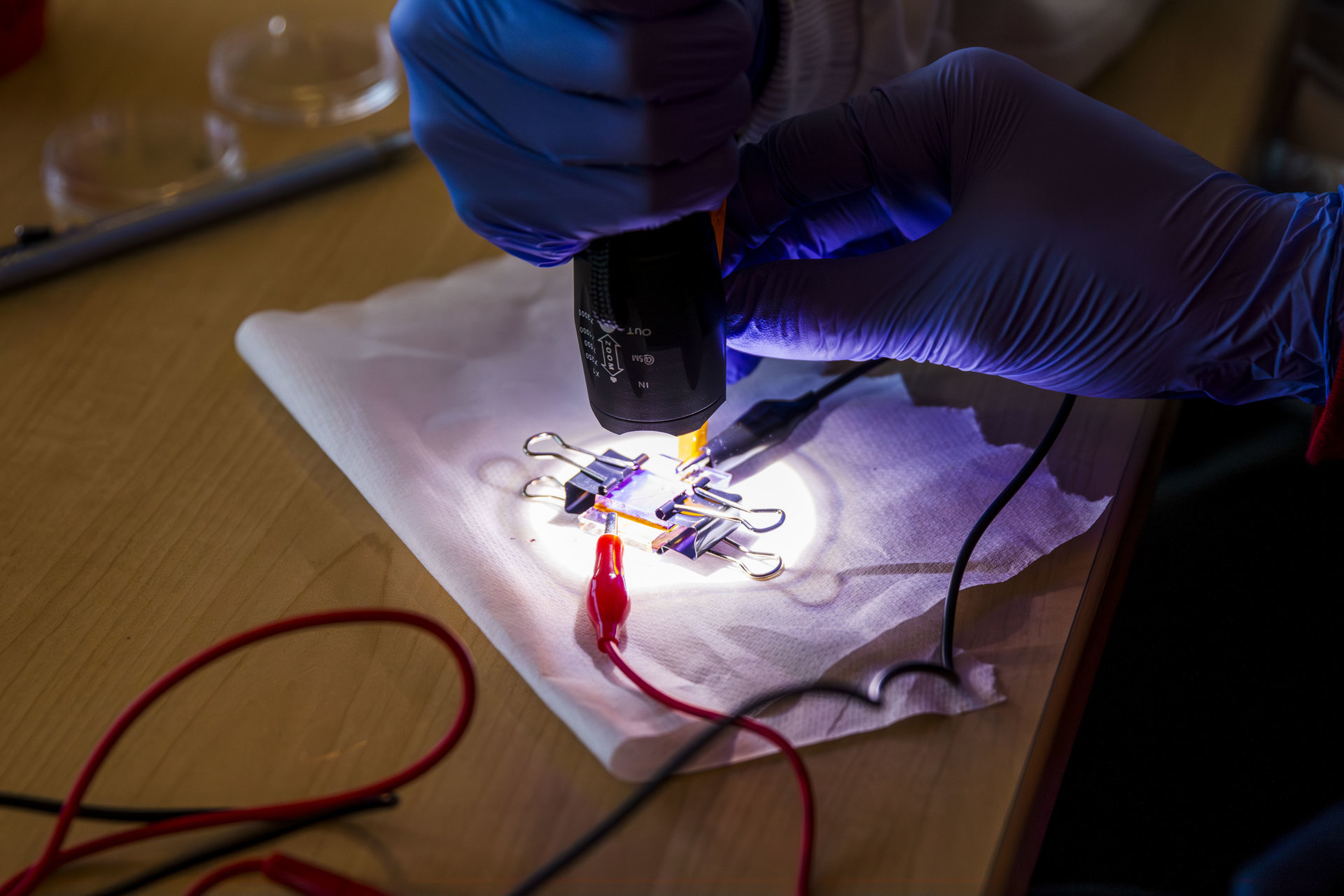 A pair of gloved hands holds a flashlight over a clear plate connected to wires.