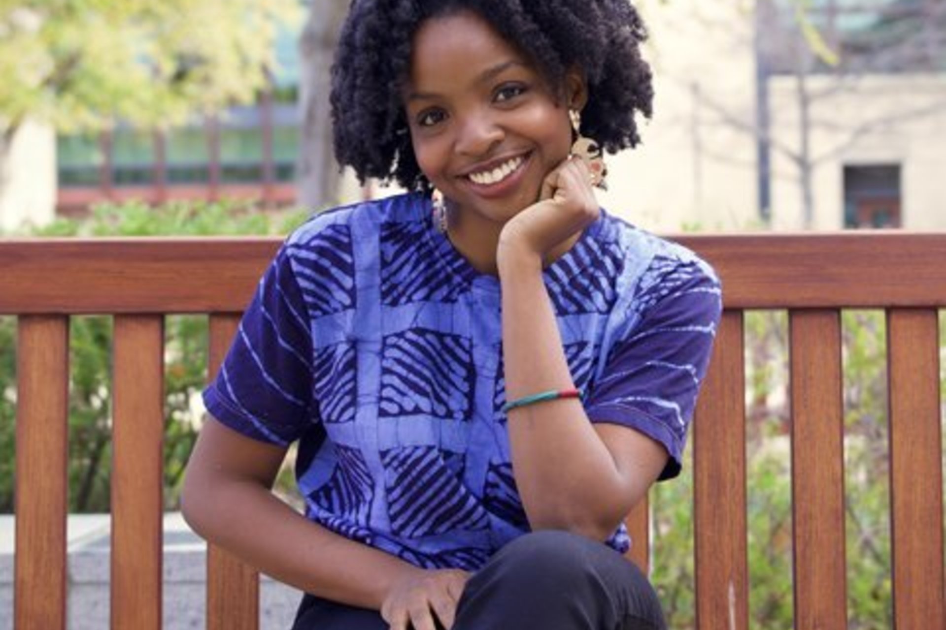 Woman in a blue shirt sits on a bench outside with chin resting on hand