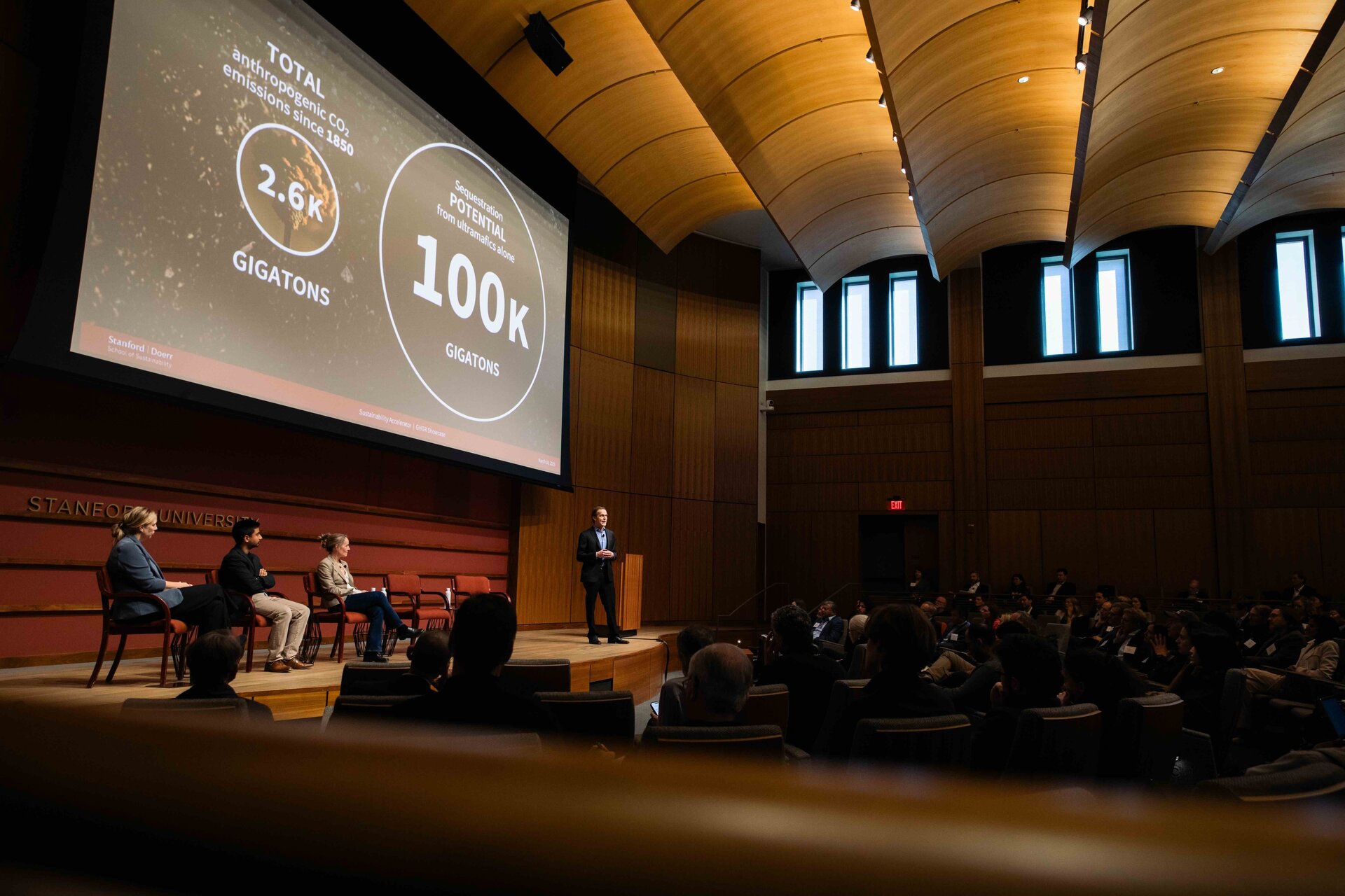 Side view of the stage with a speaker presenting while three others sit on a panel listening
