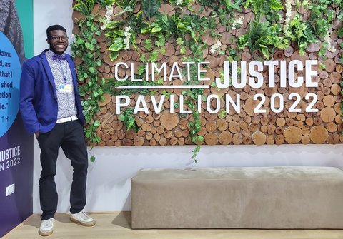 Toweh stands beside a wall with plants and the words Climate Justice Pavilion 2022