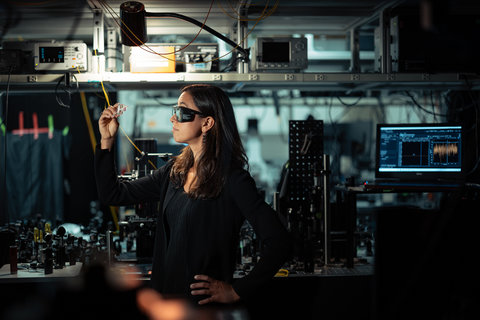 A researcher wearing safety glasses holds up a small device in an optical sensing lab.