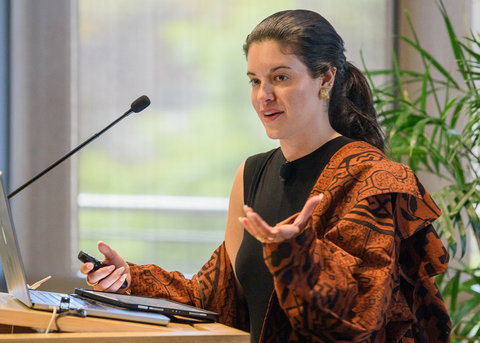 A woman wearing a rust-colored jacket with a geometric pattern gestures as she speaks into a microphone at a podium