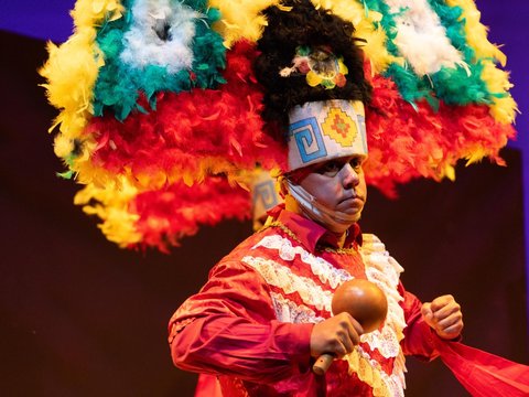 A dancer in a colorful feathered crown holding a rattle, performing La Danza de la Pluma
