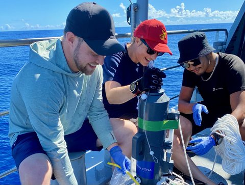 Three researchers gather around a Niskin bottle, a cylindrical device used for collecting water samples, on the deck of a research ship with the ocean in the background.