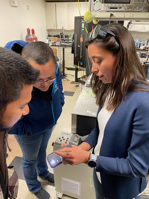 A researcher uses their iPhone to illuminate a small chip for two other researchers to see. They are standing in a lab.