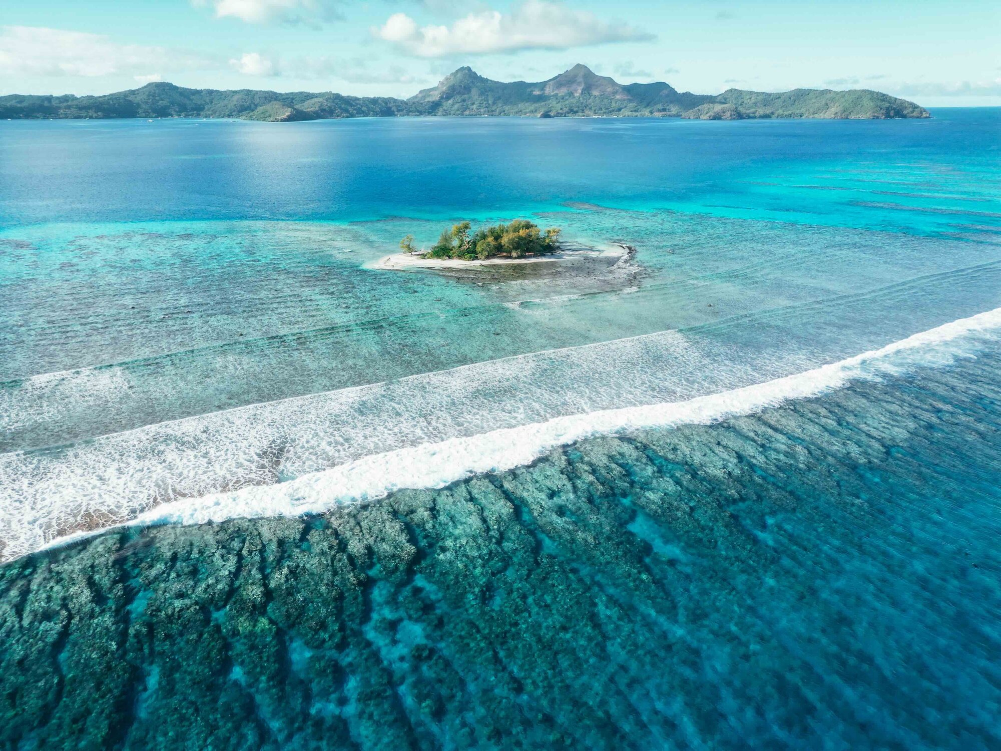 Overhead view. Small islet in clear teal water, wave rolling by. Reefs below in row formations. Island in background