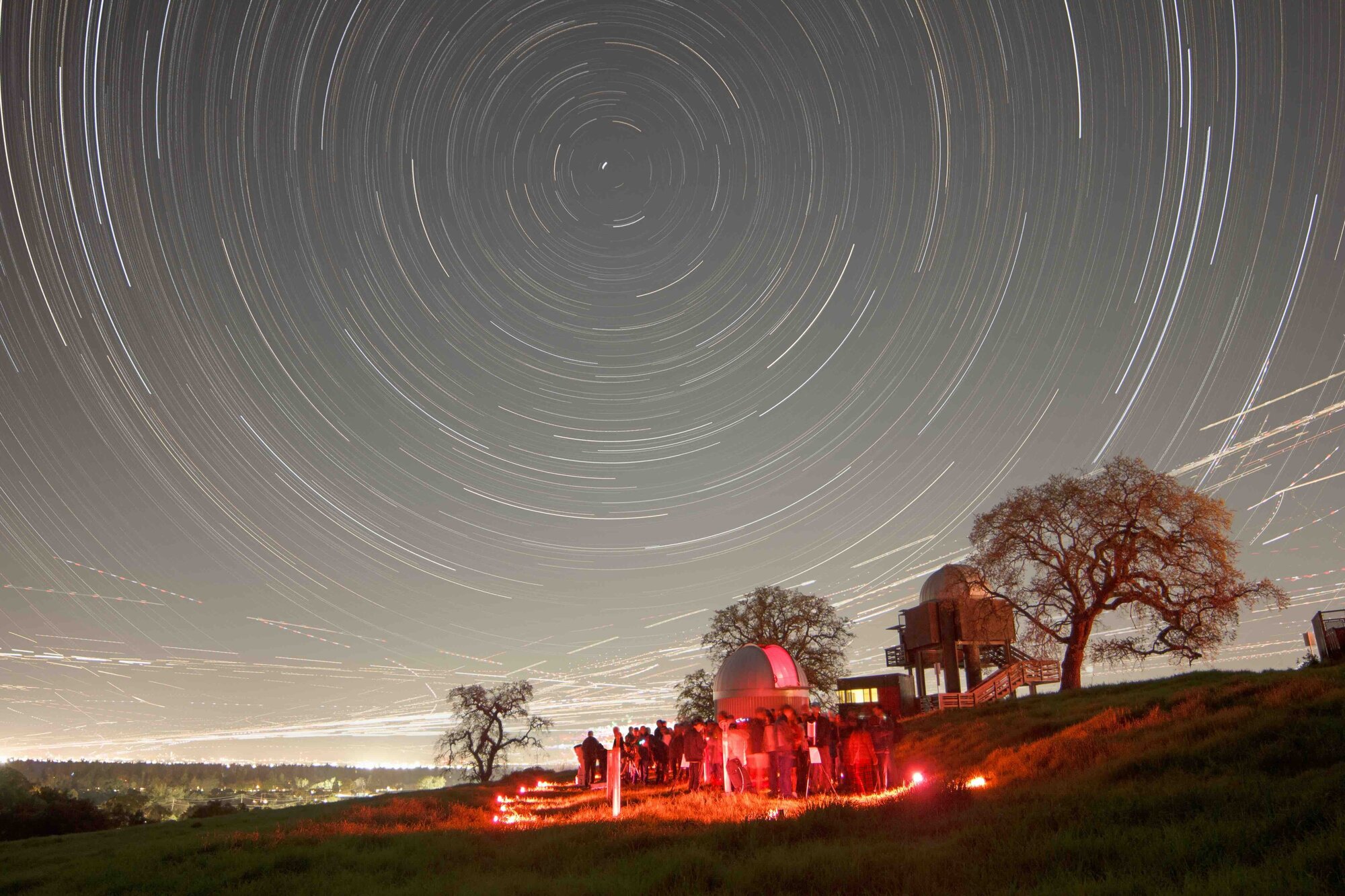 Vast spiraling streaks of white lines in gray night sky. Below, a red glow on a small crowd of people and an observatory