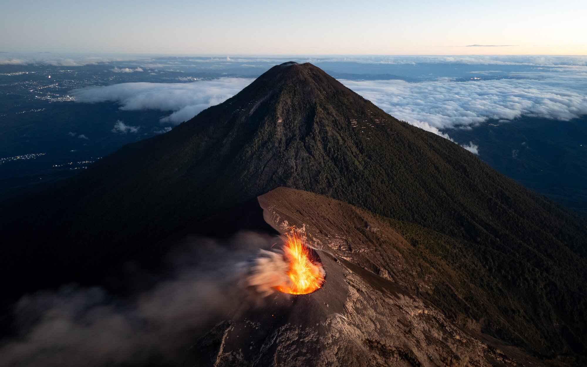 Overhead view of two volcano peaks, orange lava and smoke spewing from lower one. Morning sky, flat clouds in background