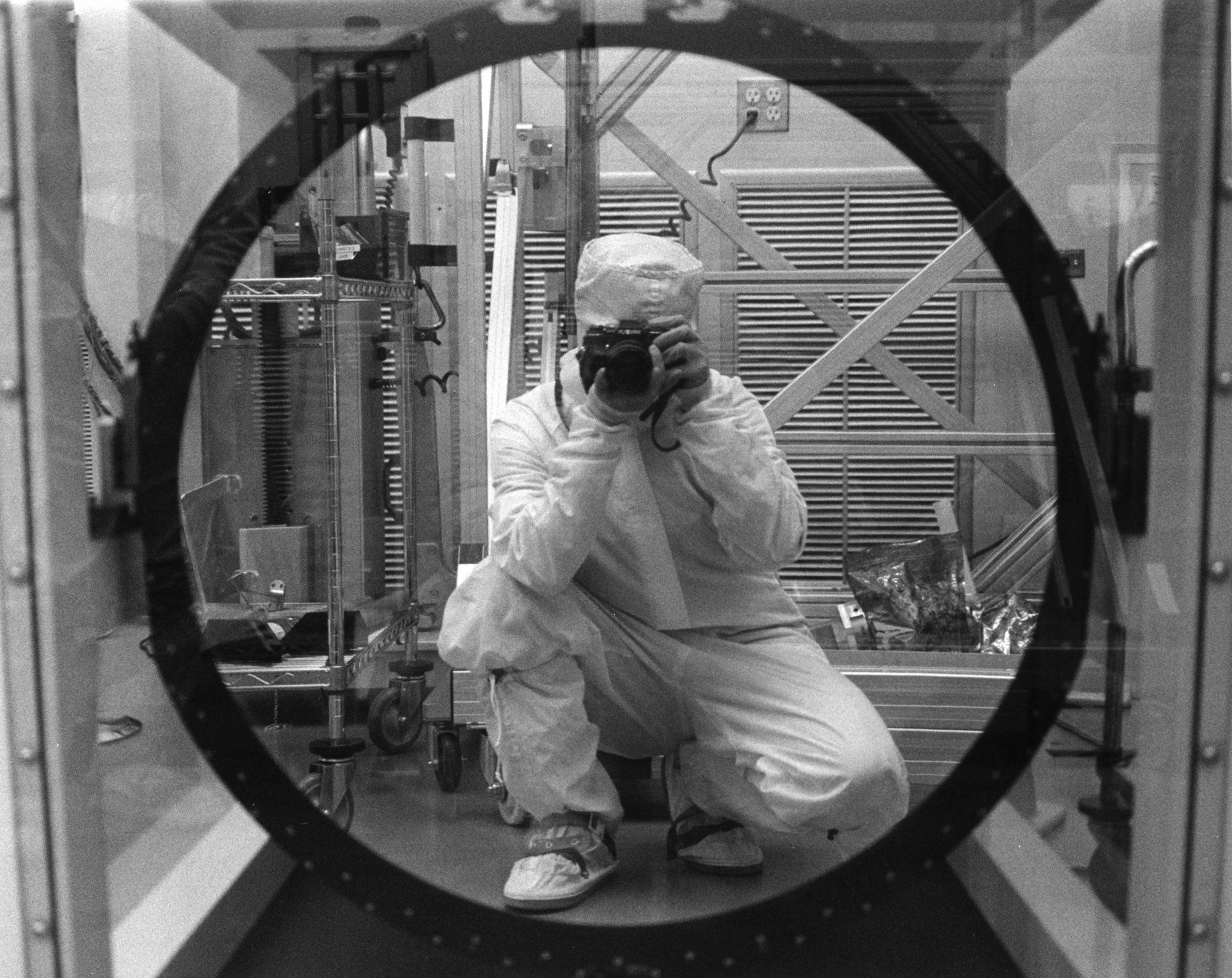 Black and white. Person in a protective lab suit takes a photo of their reflection, framed in a large circular mirror