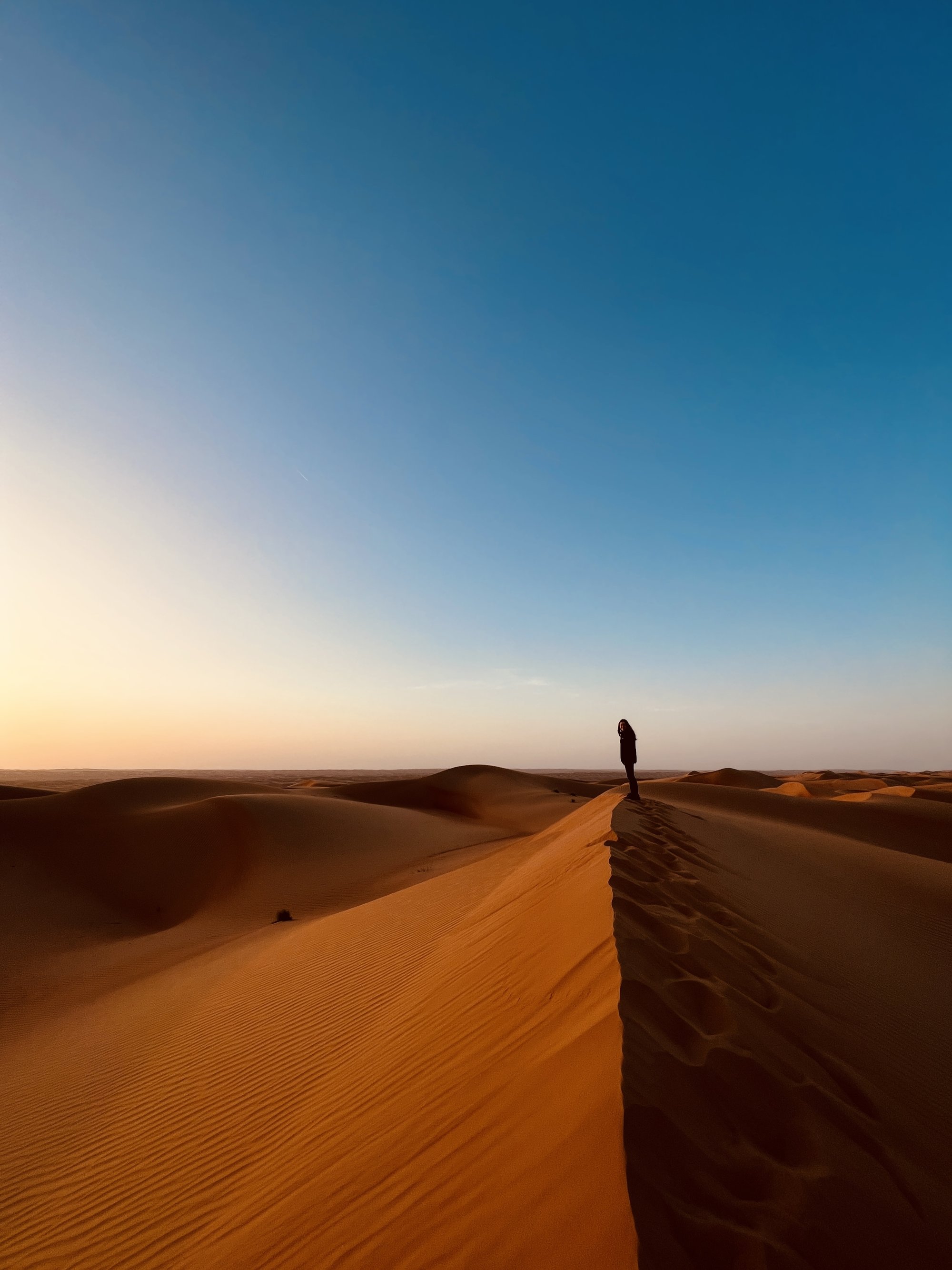 Silhouette of person standing on sand dune peak. Horizon divides bright orange dunes from deep blue sky