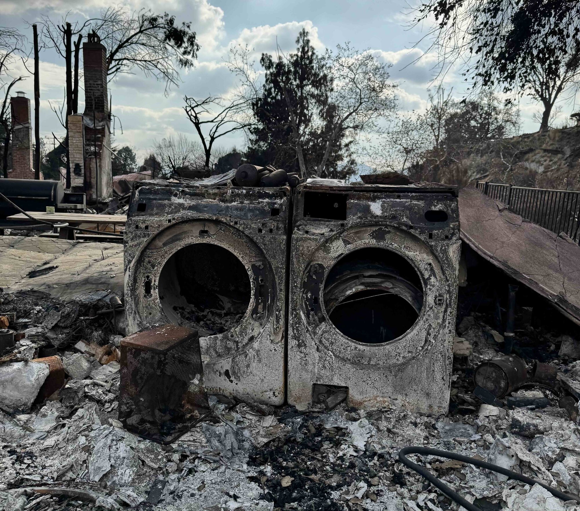 Burnt washer and dryer stand among house rubble. Light blue sky and trees in the background