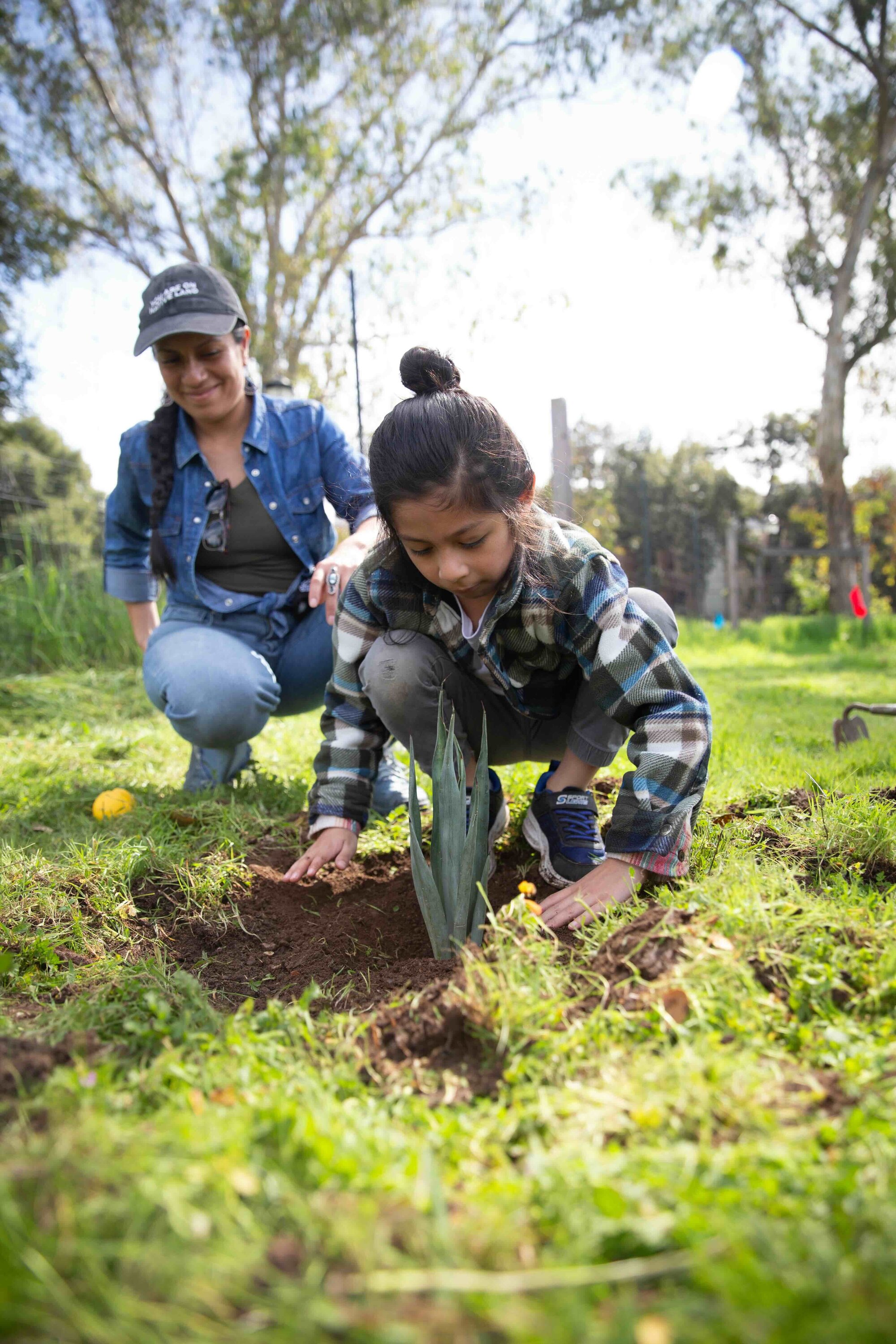 A child pats down soil around a plant in the ground while a person smiles behind