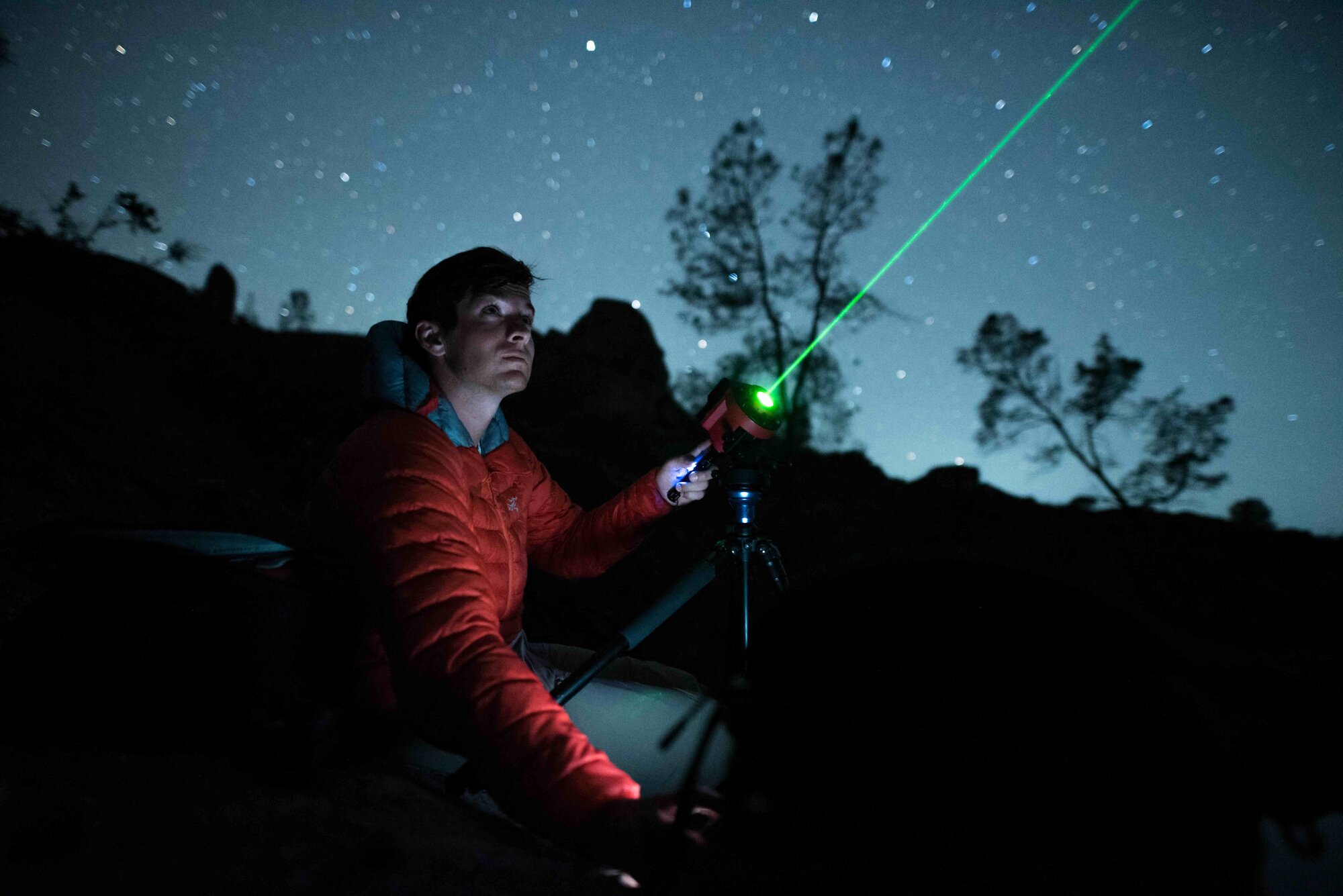 Looking up, a person aims a green laser pointing diagonally into the night sky. Background of stars and tree silhouettes