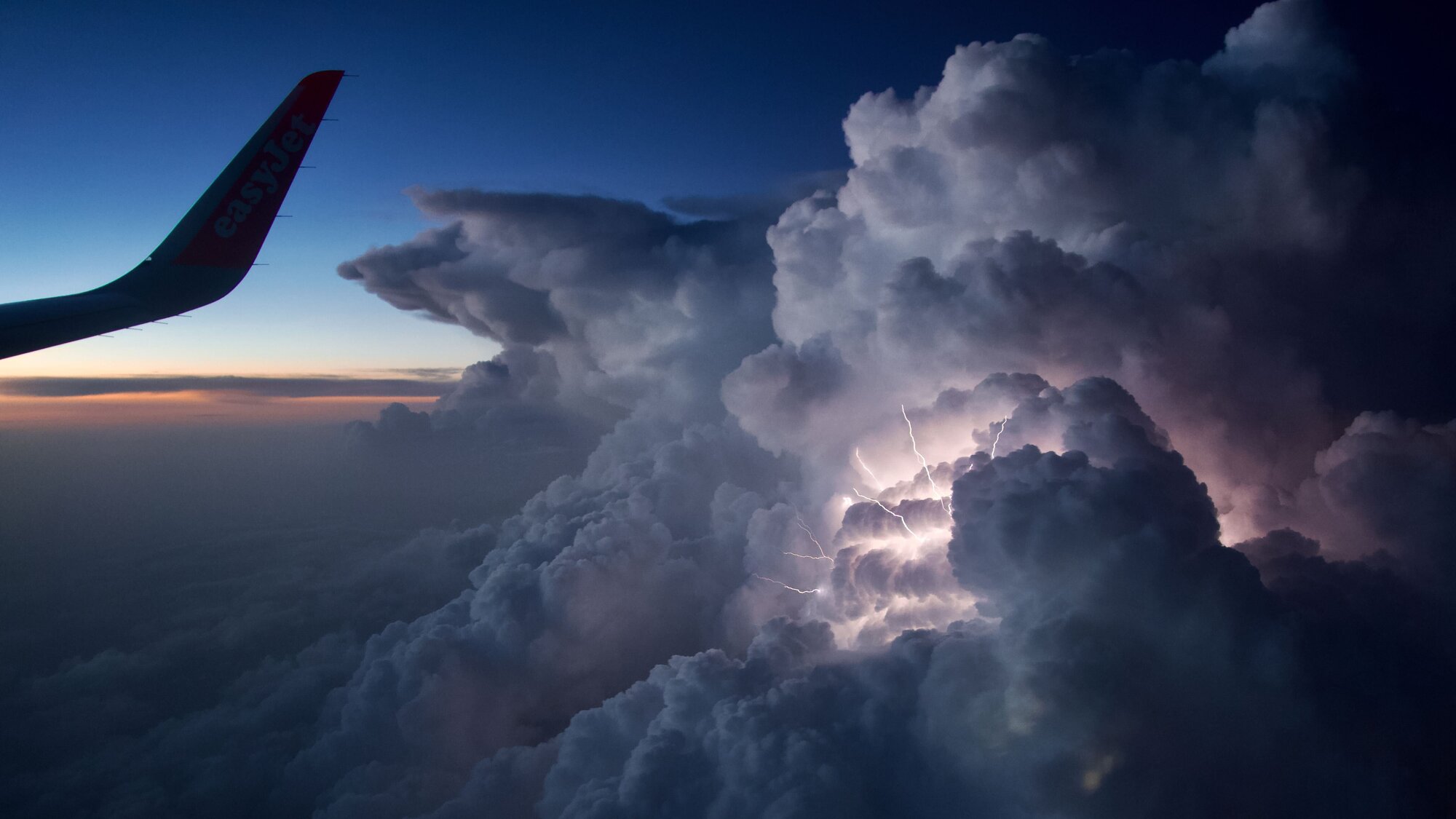 Lightning strike illuminates puffy gray and purple clouds on right. Plane wing on left and sunset in distance