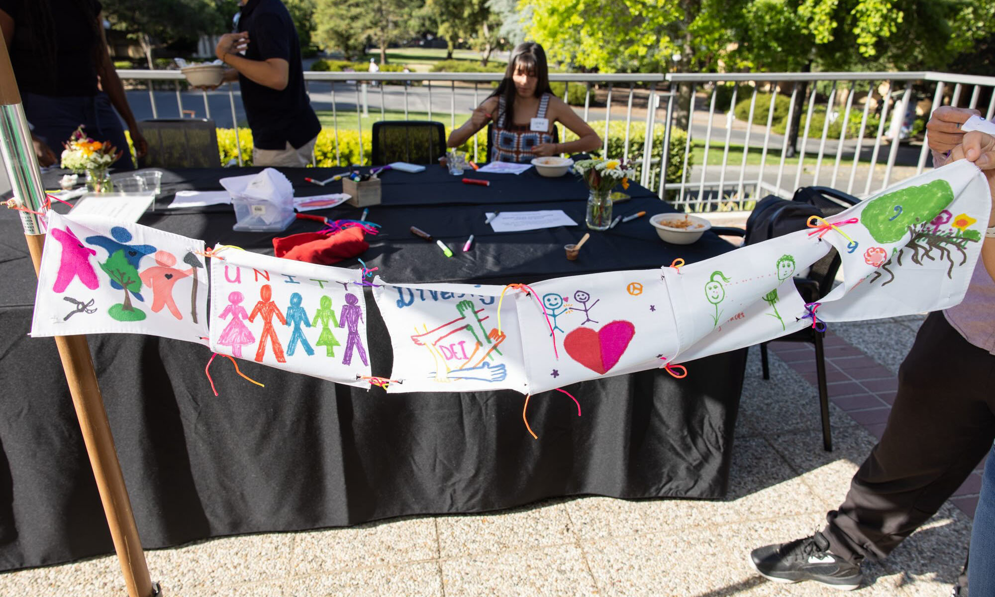 A banner of fabric squares tied together, decorated with colorful illustrations of people, trees, and diversity
