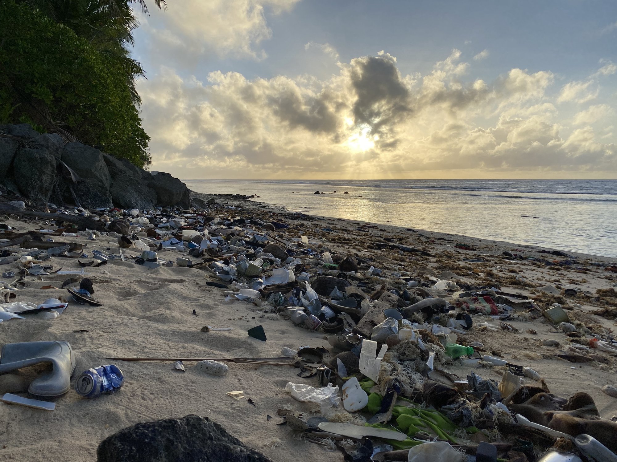 Sun rays shining through bright clouds above the ocean. In the foreground, the beach is cluttered with trash