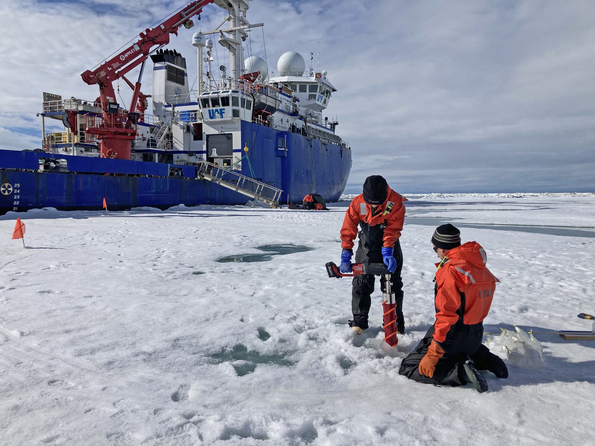 Researchers in bright orange jackets drilling into a sheet of ice with a large blue research vessel in the background
